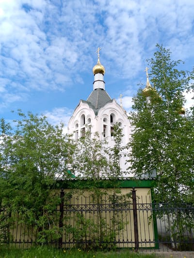 The image shows a white church with golden domes against a bright blue sky filled with scattered white clouds. The church has arched windows and crosses atop its domes. In the foreground, a black metal fence with decorative elements runs horizontally across the frame. Beyond the fence, lush green trees and foliage partially obscure the lower part of the church building. The ground in front of the fence is covered with green grass. The church is located in the city of Neryungri, Russia.