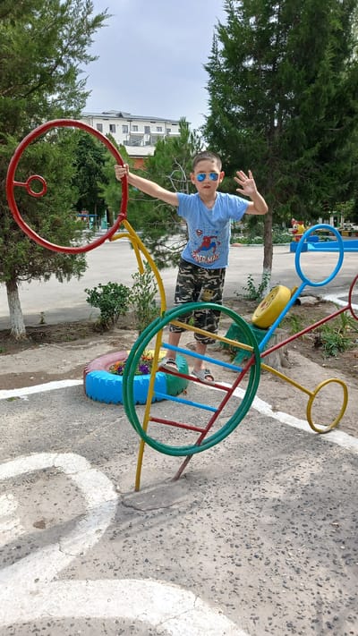 A young boy, wearing sunglasses and a blue Spider-Man t-shirt with camouflage shorts, poses playfully on a colorful playground structure in Fergana City, Uzbekistan. The structure appears to be made of repurposed materials, with bicycle wheels, tires painted in bright blues and greens, and metal bars forming its design. The boy is holding onto a red hoop, his left arm extended towards the camera with a raised hand, suggesting a moment of fun and interaction.
The setting is an outdoor playground with a concrete surface marked with white lines. Green trees, characteristic of the region, surround the area, with a multi-story building visible in the background, likely an apartment complex or school. The sky is overcast, indicating it might be a cloudy day, possibly morning or afternoon. The overall mood is cheerful and energetic, with the boy's pose and expression conveying a sense of joy. The image captures a typical scene of childhood activity in an urban environment, adapted with creative use of recycled materials.