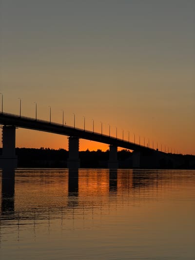 A long, modern bridge, silhouetted against a vibrant sunset, spans across a wide body of water. The sky transitions from a deep grey at the top to a warm orange and yellow glow near the horizon. The bridge, with its numerous streetlights, is a dark, imposing structure, supported by large concrete pillars. The water below reflects the fiery colors of the sunset and the dark silhouette of the bridge, creating a shimmering, distorted image. On the far bank, a dense line of trees forms a dark, unbroken horizon. The scene appears tranquil and expansive, capturing the beauty of a sunset over a bridge and river. There is no indication of human activity or specific landmarks that would identify it as being in Tarasovo, Russia.