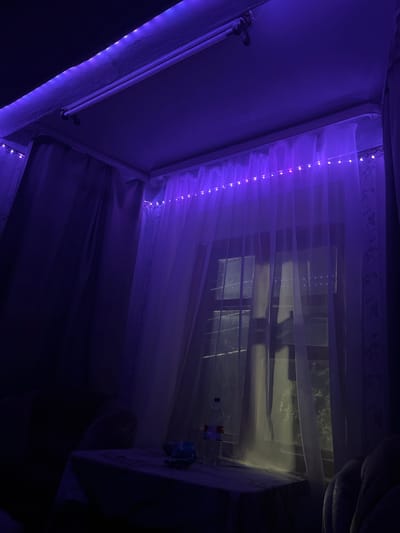 A dimly lit room is illuminated by purple LED strip lights along the ceiling and draped across the sheer curtains. A large window, partially obscured by the curtains and a subtle reflection, shows a dark, outdoor environment with glimpses of greenery, suggesting it is nighttime. In the foreground, a table covered with a patterned cloth holds a plastic bottle of water and two small dark objects. The overall atmosphere is dark and moody, with the purple lighting casting a vibrant glow on the curtains and ceiling. The location is not specified in the image or its context.