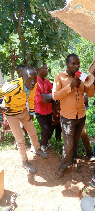 A group of men are gathered outdoors. One man in an orange shirt is holding a megaphone. Another man wears a yellow jacket and tan pants, while another is in a red shirt and dark pants. A burlap sack hangs overhead, providing partial shade. The men are standing on bare ground, some wearing sandals or shoes. Green foliage surrounds them.