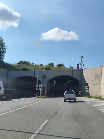 The image shows a view from a vehicle driving towards a dual tunnel entrance on a sunny day. The road in the foreground is asphalt with white lane markings. A white truck is partially visible on the left, its trailer loaded with black cylindrical objects. A silver SUV is driving ahead of the camera, approaching the tunnel.
The tunnel entrance is a large, arched structure made of concrete. There are two distinct openings, suggesting a two-lane tunnel or separate tunnels. Green lights are illuminated above the entrance of the right tunnel opening, indicating it is open. Red lights are visible above the left tunnel opening, suggesting it may be closed or for specific traffic.
Several traffic signs are present near the tunnel entrance. One sign shows a circular prohibition symbol with a horizontal bar and a green tunnel symbol below it, indicating a tunnel is ahead and possibly advising against something. Another sign has a green square with a white tunnel icon and "650M" below it, indicating the tunnel is 650 meters ahead.
The area around the tunnel is landscaped with grass and bushes on an elevated embankment. A fence runs along the top of the embankment. A cellular antenna tower is visible on the right side, mounted on top of the concrete structure. The sky is clear blue with a few white clouds. The location is identified as being in the vicinity of Golemo Buchino, Bulgaria.