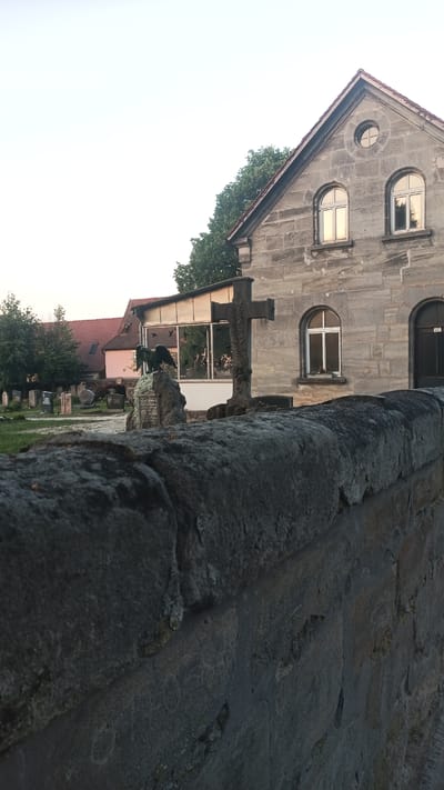 The image shows a stone wall in the foreground with a cemetery visible in the background. Behind the cemetery is a building with a light-colored stone facade and a red tiled roof. The building has arched windows and a circular window in the gable. A stone cross stands in front of the building, and a statue of an angel is on a tombstone in the cemetery. The sky is a pale, overcast color. The location is Altdorf bei Nürnberg, Germany.