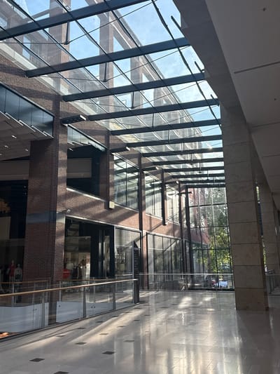 This is a wide-angle, high-angle shot of a modern atrium with a glass ceiling, likely taken in the daytime. Sunlight streams through the glass, creating strong shadows and highlights on the tiled floor. The atrium features multiple levels of a building constructed with brick and glass. Some levels have open balconies with glass railings, and some appear to be retail spaces with mannequins visible in shop windows. Trees are visible through the glass ceiling, suggesting the atrium is at or near ground level and open to the outside. The scene is bright and appears to be well-lit by natural light, implying clear or partly cloudy weather. No specific activity or event is clearly depicted, but the presence of mannequins and shop windows suggests a commercial setting, perhaps a shopping center or a large department store. There is a red "50%" sign visible in one of the shop windows. The overall impression is one of modern architecture and a lively, well-lit public space.