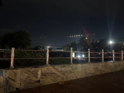 The image depicts a nighttime scene in Chennai, India. In the foreground, a polished metal railing with vertical supports runs horizontally across the frame. This railing sits atop a patterned granite wall, with a dark, wet surface below that appears to be a pathway or ground.
Beyond the railing, the scene opens up to a landscape at night. Several trees, dark and silhouetted against the sky, are visible on the left. Further in the background, a construction site is evident, dominated by a tall, red crane. A row of lights illuminates a section of the construction area, casting a bright glow into the night sky. Other buildings and structures are dimly visible in the distance, some with lights on. The sky is dark, with some faint wisps of clouds or atmospheric haze. Light beams are visible, suggesting the presence of strong, directional lighting, possibly from the construction site or streetlights. The overall impression is one of ongoing urban development in a nighttime setting. There is no visible text in the image.