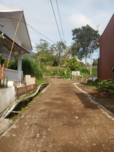 The image depicts a narrow, cracked, and stained asphalt road winding uphill between buildings and overgrown vegetation. On the left is a building with a white facade, a sign that reads "ARANAA VILLA W 42 DUNGUN BUKOR," and a wooden fence. A series of white pipes run along the base of the building. On the right is a tall, red building with an antenna visible above the trees. Lush greenery, including trees and overgrown plants, lines the road on both sides. Debris is scattered along the roadside. Power lines stretch across the sky. The location is inferred to be in Ein Chay Hnit Se, Myanmar, based on the sign. There are no people visible.