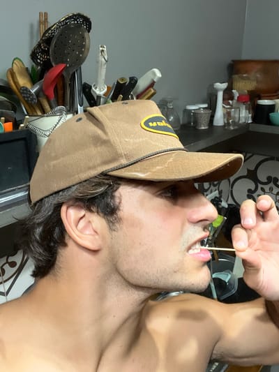 A shirtless man, appearing to be in an indoor kitchen or utility area in Avellaneda, Argentina, is depicted in a portrait-style shot. The image is rotated 90 degrees clockwise. He has dark, curly hair and stubble/beard on his face, and his head is tilted upwards with his mouth slightly open. He is wearing a brown cap with a yellow patch on the front, where the letters "an" are partially visible in black text.
The man's right hand is visible, holding a wooden toothpick which he has placed inside his mouth, touching his tongue. His attention appears focused on the toothpick. The man's muscular shoulders and neck are visible on the right side of the frame.
The background shows a cluttered shelf or counter filled with various kitchen items. These include numerous utensils such as a slotted spoon, a red ladle, knives, whisks, and other implements, along with several jars, bottles, and a scrubbing brush. Behind the man's head, a decorative dark brown or black pattern on a light-colored tiled wall is visible. The lighting is bright and even, highlighting the man and the nearby objects.