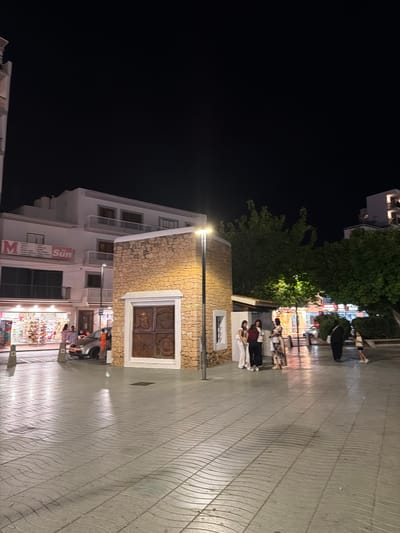 The image depicts a street scene at night in Sant Antoni De Portmany, Spain. A paved walkway with a wave-like pattern occupies the foreground. On the left side of the image, a multi-story white building with balconies and shops on the ground floor is visible. One shop has a sign that reads "M" and "Sun." A silver car is parked in front of the building.

In the center of the scene, a small, rectangular building constructed of rough-hewn stone with a decorative metal door stands prominently. A street lamp illuminates the area. A group of four young women are standing near the stone building, appearing to be in conversation. Further down the street, several other people are walking, including a person dressed in black and a young girl. The background features more buildings and lush green trees, with the dark night sky above. The overall impression is of a lively public square at night.