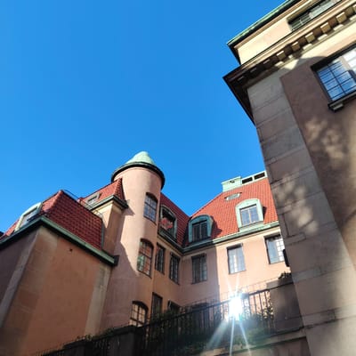 The image captures a worm's-eye view of residential buildings against a clear, vibrant blue sky in Stockholm, Sweden. The primary structure on the left features a warm, light reddish-brown (salmon-toned) stucco facade, characterized by multiple windows and a prominent round tower capped with a patinated green copper dome. Its roofs are steeply pitched and covered with red terracotta tiles, interspersed with several dormer windows featuring light green copper flashing. Green copper gutters are also visible along the eaves.

To the right, a contrasting building appears partially, revealing a textured, darker stone facade with pronounced architectural details, including horizontal bands and corbel-like elements below a green-edged roofline. This structure is largely cast in shadow, with strong sunlight illuminating the main building. A brilliant sun flare emanates from the lower right quadrant of the image, suggesting a low sun angle and reflecting off unseen surfaces, casting long, distinct shadows on the building facades. In the lower foreground, a dark metal fence draped with green ivy-like foliage partially obscures the base of the structures. No individuals are present. The architectural style, with its blend of National Romantic and Art Nouveau influences, is characteristic of buildings constructed in the late 19th to early 20th century in Stockholm, specifically consistent with the residential architecture found in the **Östermalm district**, such as along Tyskbagargatan or similar surrounding streets.