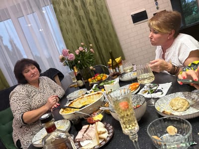 This is a medium shot, taken indoors at night, of two women sitting at a dining table laden with food and drinks. The table is black and speckled, and the background features a white brick wall and green curtains with a leaf pattern.
The woman on the left has dark, curly hair and is wearing a beige patterned blouse. She is looking towards the right of the frame with a neutral expression. The woman on the right has short, blonde hair and is wearing a white textured blouse. She is looking down at her hands, which are reaching for a smartphone, with a contemplative expression.
The table is covered with various dishes, including a casserole, a plate of sliced meat and bread, and a bowl of fruit. There are also several glasses, some filled with a pale yellow liquid, presumably champagne or white wine, and a bottle of what appears to be alcohol in the foreground. The overall scene suggests a dinner gathering or celebration. The image was taken in Malmyzh, Russia. There is no visible text in the image.