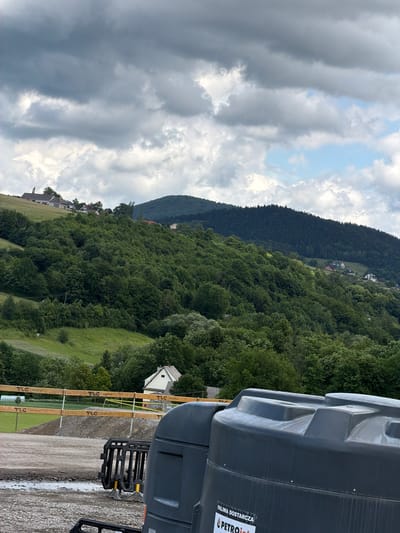 In the city of Stróża, Poland, a construction site is visible in the foreground, featuring two large, dark grey plastic water tanks. One tank is prominently positioned on the right side of the frame, with a label that reads "PALIWA DOSTARCZA" and "PETROJET." To the left of the tanks, a black plastic barrier is partially visible. The construction area is covered in gravel and dirt, with some puddles indicating recent rain. A yellow caution tape is strung across a wooden fence constructed with posts labeled "TLC" and "www.tlc.eu." Beyond the construction site, a grassy hill slopes upwards, dotted with trees. Further in the background, a lush green forest covers a mountain. Several houses are scattered across the hillside and atop the mountain, with one prominent white house visible on the left side of the hill. The sky above is filled with dramatic, grey clouds, with patches of blue sky peeking through.