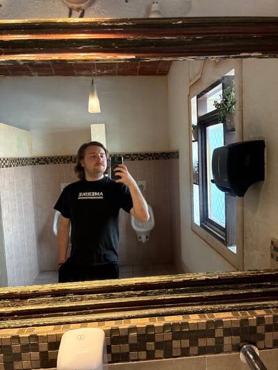 A man with brown hair and a beard, wearing a black shirt with the word "AMERIKA" printed on it, takes a selfie in a bathroom mirror. The bathroom has tan tiled walls, a toilet, a hand dryer, and a tissue dispenser. Above the man in the mirror is a window with plants visible outside. The mirror has an ornate wooden frame. The location is potentially in Tepeji de Ocampo, Mexico.