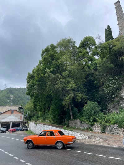 An exterior, medium shot captures a bright orange sedan driving on a wet asphalt road under an overcast sky in Abaata, Abkhazia. The car is positioned in the right foreground, facing right, with its driver visible inside. Behind the car, a stone wall with a decorative pattern runs along the side of the road, separating it from a steep, tree-covered embankment. A light-colored building, possibly a workshop or garage, is situated on the left side of the road, with several other vehicles parked in front of it. The background is dominated by lush green trees and a distant, hazy hillside.