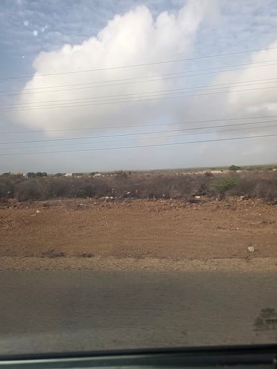 The image shows a view of a landscape, possibly taken from inside a moving vehicle. In the foreground, a section of the vehicle's interior is visible on the right. The landscape consists of a dirt or gravel area next to a line of vegetation. Beyond the vegetation, there are buildings in the distance, suggesting a small town or village. In the sky, there are power lines running parallel to the horizon and the road. To the left of the power lines, there are fluffy white clouds. The location appears to be El Dátil, Venezuela.