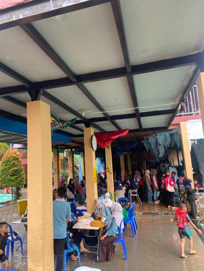 The image shows a covered outdoor dining area in Kecamatan Tanggulangin, Indonesia (latitude: -7.497, longitude: 112.684). The area is under a roof supported by thick, yellow pillars.  The roof is made of metal and has a white painted ceiling with multiple beams crossing at 90-degree angles. There is a group of people gathered under the cover, many of them are sitting and eating at tables with blue chairs. A single child in a red life vest is splashing in a puddle. The walls of the building are brightly colored and there are a few blue and red paper decorations hanging from the ceiling. The ground is made of a mixture of brown and beige tiles, which are wet and reflect some of the light. The area is empty except for the people eating and the child in the puddle. There are a few palm trees and other green plants visible in the background, along with a blue tiled pool.