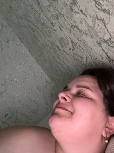 The image shows a close-up of a woman's face, turned to her right with her eyes closed and a slight smile. She has dark hair and is wearing a pearl earring. The background is a patterned surface that appears to be a countertop or wall, with grey and white marbling. A small electrical outlet is visible in the lower right corner of the image. The lighting suggests it is daytime, possibly indoors. The woman appears relaxed and content. There is no visible text in the image.