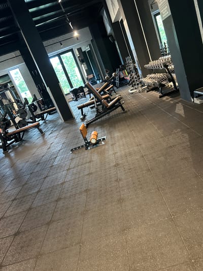 The image shows an interior view of a gym with weightlifting equipment. The floor is covered with dark gray, possibly rubberized, tiles. Weight racks filled with dumbbells and other equipment are positioned along the left side of the frame, near large windows that offer a view to the outside. The equipment is mostly black with some brown leather padding. The origin or specific location in Unknown, Romania, is not discernible from the image. There are no people visible in the picture.