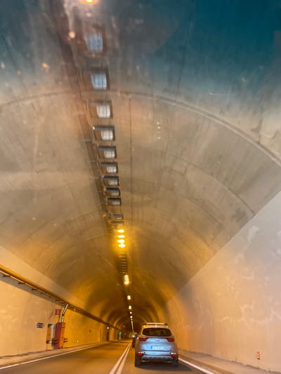 The image shows the interior of a tunnel. The tunnel walls and ceiling are made of concrete. Lights are visible along the ceiling, casting a warm glow. A silver SUV with a license plate that reads "GL 12 SEK" is driving through the tunnel. The tunnel appears to be located in Komotini, Greece, a city known for its tunnel infrastructure.