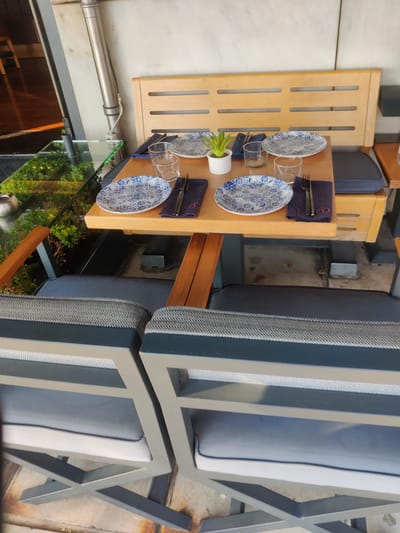 A dining table is set for two in an outdoor seating area. The table is made of light-colored wood and has a light wood bench with cushions behind it. Two place settings are visible, each with a blue and white patterned plate, a glass, and a dark blue napkin with cutlery. A small potted plant sits on the table between the two settings. The chairs in the foreground are also cushioned with dark blue and grey fabric. The background shows a wall and a glimpse of a glass-enclosed area with greenery, possibly an indoor plant display. The scene appears to be in an urban setting, likely a restaurant or cafe.