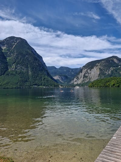 In Obertraun, Austria, a scenic view of Hallstätter See, or Lake Hallstatt, unfolds under a blue sky dotted with white clouds. The foreground features a wooden pier on the right, partially submerged in the clear, shallow water. The lakebed is visible, covered with sand and pebbles. Several individuals are engaged in water activities: two people are seen on stand-up paddleboards in the middle distance, and at least two more are swimming in the lake. The middle ground showcases the vast expanse of the lake, reflecting the surrounding environment. In the background, the imposing, tree-covered mountains of the Dachstein range frame the scene, with a small town or village nestled along the shoreline. A white boat is visible on the water near the village. The overall atmosphere is tranquil and recreational.