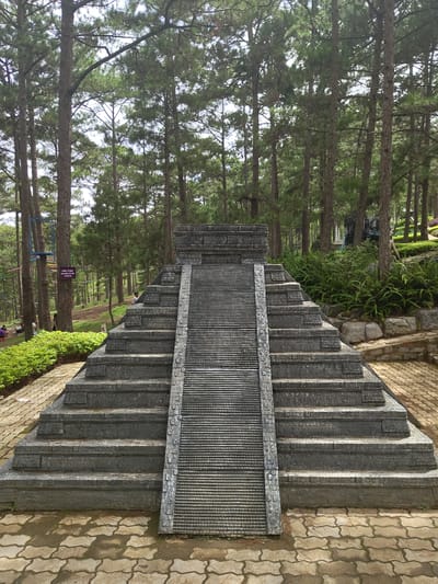 The image depicts a miniature replica of a Mayan pyramid, constructed from gray stone, situated within a park-like setting. The pyramid has a central staircase leading to a flat top. Surrounding the pyramid is a paved pathway made of interlocking bricks, bordered by lush green bushes. Tall pine trees dominate the background, indicating an outdoor environment. In the distance, a person is visible walking on a slope. A purple sign is attached to a tree on the left side of the frame, and some recreational structures, possibly part of a playground or zip-line course, are also present in the background. The scene appears to be a tourist attraction in Lam Vien - Da Lat, Vietnam, characterized by its natural surroundings and man-made monument.