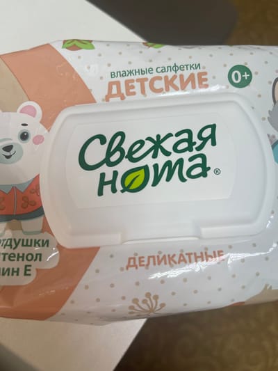 The image shows a package of wet wipes for babies. The packaging is primarily white and peach-colored, with illustrations of a cartoon bear and other decorative elements. The brand name "Свежая нота" (Fresh Note) is prominently displayed in green lettering on a white lid. The text "ВЛАЖНЫЕ САЛФЕТКИ" (WET WIPES) and "ДЕТСКИЕ" (CHILDREN'S) are also visible in Russian. There is a "0+" marking indicating suitability for newborns.
The image does not provide any information about the location in the city of Nefteyugansk, Russia.