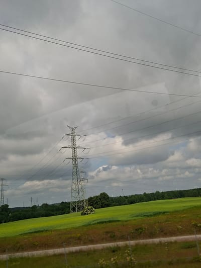 The image captures a scenic view of a rural landscape with power lines stretching across a cloudy sky. In the foreground, a vibrant green field slopes gently, bordered by a strip of reddish-brown vegetation and a fence. Several tall, metal electricity pylons are visible, the closest one prominently positioned on the right side of the frame. In the background, a dense line of trees forms the horizon, with wind turbines appearing in the distance. The sky is dominated by heavy, grey clouds, creating a somewhat dramatic atmosphere. The image does not contain any people or indicate any specific actions or interactions. The overall scene is one of natural beauty enhanced by industrial infrastructure, consistent with the countryside. The location is not specified as Suwałki, Poland.