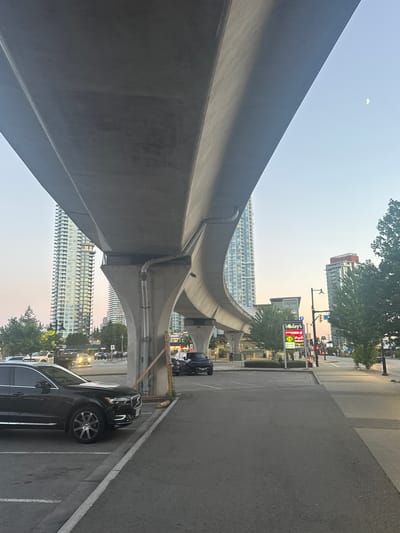This image depicts an urban scene in Coquitlam, Canada, likely near Lougheed Highway, characterized by elevated transit infrastructure and surrounding city buildings. In the foreground, a dark-colored SUV is parked in a paved lot next to a concrete curb. Beyond the parking area, a wide asphalt road is visible, with several vehicles in motion and stationary. To the left, a tall, modern high-rise building with numerous windows dominates the skyline, reflecting the ambient light. Further back, another similar high-rise is partially obscured by trees and the elevated concrete structure.

The dominant feature of the scene is the massive, curving concrete structure of an elevated transit line, casting a shadow over the foreground. This structure is supported by large, angled concrete pillars. On the right side of the road, a multi-story building with a red top is visible, along with streetlights and traffic signals. A pedestrian sidewalk runs alongside the road. In the sky, a crescent moon is faintly visible against the pale blue evening sky. Signs for businesses such as "Bosley's," "Shop Metal," and "Thrift Optical" are mounted on a pylon near the road. Trees are scattered throughout the scene, with dense foliage on the right side. The overall lighting suggests either early morning or late evening.