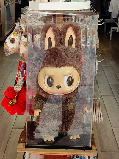 This image captures an indoor retail environment in Sa Pa, Vietnam. Dominating the foreground is a large, brown, shaggy-furred character figure with a light-toned, humanoid face, large eyes featuring white sclera and starburst-patterned pupils, a small black nose, and prominent ears. The figure has visible light-toned hands and three-toed feet, and a circular metallic tag is affixed to its chest. It is enclosed within a clear, rectangular acrylic display case with a black base, which rests upon a light-colored wooden stand.
To the left of the display case, multiple transparent plastic bags, each containing what appear to be food items (possibly snacks with a white coating and brown pieces), hang from hooks. Below these bags, several red plush items, some accented with yellow details and equipped with attached tags, are visible. To the right of the display case, a vertical panel features a series of silver metal hooks, from which various packaged goods are suspended, though specific details of these items are indistinct. The background reveals additional shelving units stocked with merchandise and racks holding light-colored clothing. The floor is composed of light-toned wooden planks.