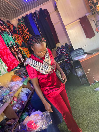 An adult female is depicted standing within a retail clothing establishment in Samaru, Nigeria. She has braided hair with red accents and wears a red velvet short-sleeved tunic with embellished detailing on the chest, along with matching red velvet trousers. A plaid-patterned shoulder bag with a black strap and a small black purse are worn across her body. Her gaze is directed downwards, and her right hand holds a clear plastic bag containing unidentified items.
The environment features a corrugated metal ceiling and light-colored paneled walls. To the left, multi-tiered shelves display numerous items, presumably clothing or textiles, individually packed in clear plastic bags of various colors. Along the back wall, several garments, including dresses and kaftans in diverse patterns (e.g., floral, abstract, animal print) and solid colors (e.g., green, red, orange, blue, black), are hanging on display racks. To the right, a wooden counter is visible, on which rests a black bag and a dark, flat object resembling a laptop or notebook. The floor is covered with green artificial turf. Overhead lighting fixtures are present, with a blue light visible near the ceiling.