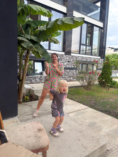 In Baranovka, Russia, a woman and a young child are photographed outdoors in front of a modern building. The woman, with dark hair, wears a pink and green patterned dress and light-colored sneakers. She stands with her arms raised, her hands in fists, and a wide smile on her face. To her right, a blond-haired child in a gray t-shirt and shorts, with visible socks and sandals, stands with an open-mouthed expression of surprise or excitement. The child's right arm is extended, and their left arm is bent. Behind them, a large banana tree with broad leaves is visible, with the building featuring dark patterned stone accents and large windows. A rose bush with red blooms and a conical evergreen tree are also present in the background, along with a grassy area and a concrete walkway. A portion of a wooden chair with a worn fabric seat is in the foreground on the left.