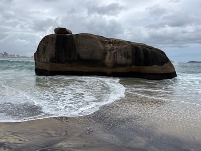 This photograph captures a rugged coastal scene in Rio de Janeiro, Brazil, dominated by a massive, dark rock formation at the edge of the ocean. The sky is overcast with heavy grey clouds, suggesting a cloudy or stormy day, but it is clearly daytime.
The primary subject is a very large, dark grey rock, appearing like a huge boulder, situated in the shallow waters where the waves meet the shore. Its lower section, frequently submerged, appears darker and more saturated, possibly from being wet or a tidal mark. On top of this main rock, a smaller, similarly dark rock rests, and a tiny white bird, likely a seagull, is perched atop the smaller rock, looking towards the distant city.
The ocean waves crash around the base of the large rock and gently roll onto the sandy shore in the foreground. The water is a deep grey-green, with frothy white foam from the breaking waves creating intricate patterns on the wet sand. The sand itself shows variations in tone, from lighter tan to darker, almost black, likely due to moisture content and mineral composition.
In the distant background, a cityscape of high-rise buildings lines the horizon, partially obscured by the overcast sky, indicating an urban presence near this natural landmark. To the far right, a faint outline of another landmass or island can be seen across the water.
There is no discernible human activity or visible text in the image. The overall mood is one of natural grandeur and a slightly muted palette due to the cloudy weather.