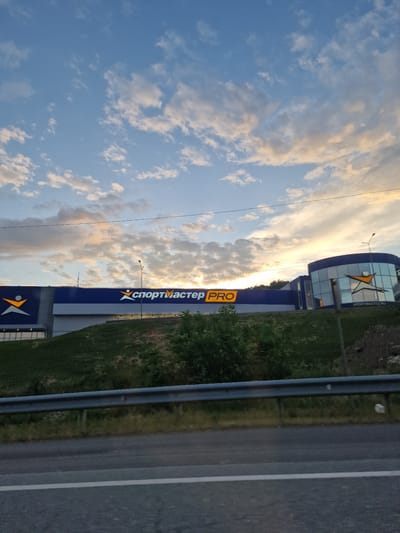 The image captures a wide shot of a Sportmaster PRO store in Goryachevodsky, Russia, under a cloudy sky with hints of sunset colors. The building features a prominent blue facade with large white lettering spelling out "СПОРТМАСТЕР PRO". The store's logo, a stylized orange figure with outstretched arms, is visible on the left side of the building. To the right, a section of the building is rounded with glass windows, also displaying the Sportmaster logo and the website "SPORTMASTER.RU". The store is situated on a grassy incline, with a metal guardrail and a paved road in the foreground. A street lamp is visible on the right side of the building. The overall impression is that of a commercial establishment on the outskirts of a town or city, seen from a road.