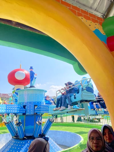 This image captures an outdoor amusement park scene in the city of Batu, Indonesia, under a clear blue sky with minimal scattered white clouds. The upper portion of the frame is spanned by a large, curved archway structure, painted vibrant yellow on top, transitioning to dark green, and then a lighter green edge against the sky.

In the lower right foreground, two women wearing hijabs are partially visible. The woman closer to the viewer wears glasses and a light purple hijab, while the woman behind her wears a dark blue hijab. Both appear to be looking towards the camera.

The central subject is a colorful amusement park ride. It features a prominent blue central column decorated with numerous white circular studs. Radiating from this column are several light blue seats or gondolas, each equipped with a safety bar. Multiple individuals are seated in these carriages; on the left side, two occupants wear dark blue tops and denim jeans, and on the right, another person in a dark top is visible.

Mounted atop the ride's central structure are two large decorative figures: a red mushroom with a white circular spot on its cap, and a standing male figure in a dark blue top hat, suit, and holding an umbrella, facing away from the viewer.

The background beyond the ride includes a meticulously manicured green lawn, partially bordered by a metal fence. A white utility vehicle is visible behind this fence. Further in the distance, buildings with red-tiled roofs are discernible. Colorful flowerbeds are distributed throughout the scene, and a section of a decorated wall, featuring an orange brick pattern and a mural resembling yellow frosting with sprinkles, is visible in the upper right background.