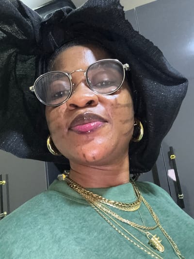 In Ajara, Nigeria, a woman is shown in a close-up, upper-body photograph. She is wearing a black headwrap and round, silver-rimmed glasses. Her lips are glossy and a vibrant pink. She has on a green sweatshirt and is adorned with multiple gold necklaces, including a thick chain and thinner layers with a pendant shaped like a capsule. Large gold hoop earrings are visible on each ear. The background appears to be an indoor setting with grey cabinets and doors.