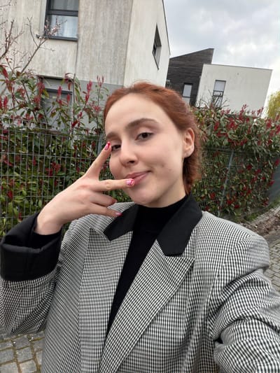 The image portrays a young female subject standing outdoors in what is identified as Eschau, France, beneath an overcast sky. The subject, possessing reddish-brown hair, faces the camera with a slight smile. Her left hand is raised, forming a V-sign with extended index and middle fingers, while the ring and pinky fingers are curled. Her visible fingernails exhibit a white base polish adorned with red and pink curvilinear patterns. She is dressed in a black high-necked garment beneath a black and white checkered blazer, which features solid black lapels and rolled-up cuffs revealing a black lining. A small silver stud earring is visible in her right earlobe.
The background comprises modern residential architecture, consisting of at least three buildings with light-colored stucco facades. One building displays a prominent dark, angular roof section and a diamond-shaped window. A metal fence separates the subject from a dense hedge characterized by green foliage with vibrant red new growth. Bare tree branches with small buds are visible to the far left, and a weeping tree with yellowish leaves is partially visible on the far right. The immediate foreground shows cobblestone paving.