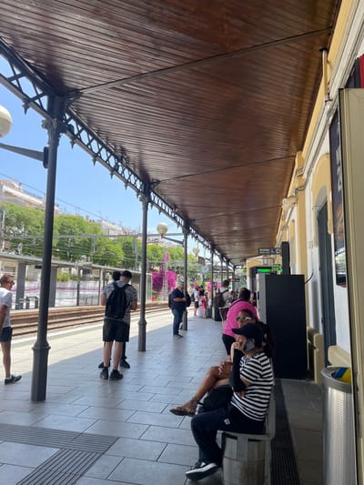 This image depicts a train station platform in Sitges, Spain, likely on a sunny day. Several people are visible, some sitting and others standing, appearing to wait for a train. The platform is covered by a wooden roof supported by ornate metalwork. The background shows train tracks, lush green trees, and buildings.
In the foreground, a person wearing a black and white striped shirt and black pants is seated, holding a mobile phone. To their left, another person in a pink top and sunglasses is also seated. Further down the platform, people are spaced out, adhering to social distancing guidelines, as indicated by the empty spaces between them. One person is wearing a mask.
Visible text includes signage with "Sortida" (Exit) and "Salida" (Exit) in Spanish, along with an accessibility symbol. There is also a green illuminated sign with Spanish text, likely indicating train information. The overall atmosphere appears relaxed, with people patiently waiting. The bright sunlight suggests it is daytime.