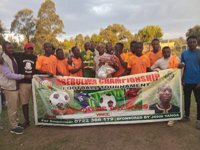 An outdoor photograph captured in Chevaywa ward, Kenya, shows approximately 18 individuals, primarily young adult males, posing behind a horizontal banner on a grassy field. The banner, central to the image, reads "ABEBUWA CHAMPIONSHIP FOOTBALL TOURNAMENT" and "NURTURING TALENTS," displaying images of footballs, football players, and a portrait of "JOHN YANGA," identified as the sponsor.
On the far left, a male in a light-colored jacket and khaki shorts stands next to another male wearing a black t-shirt printed with "OPPOSITION LEADER" and a red cap. The majority of the group, consisting of about 14 males, are dressed in matching orange football jerseys and dark shorts. One male in a green shirt holds two gold-colored trophies. Another male in a light-colored top holds several white footballs and a red and white bundle. To the far right, a male wears a blue t-shirt, dark trousers, and carries a dark backpack.
The background features numerous trees with light-colored trunks and sparse foliage, indicating an open field or park environment. Faint, light-colored structures are discernible in the distance on the right. The sky is overcast, producing soft, diffused lighting, suggesting late afternoon or early evening. The ground is covered with dry grass and dirt.