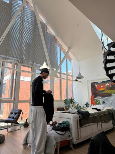 This image depicts an interior scene, identified as being within the city of Saint-Denis, France, consistent with a residential loft or apartment. A lean male individual, wearing a black long-sleeved shirt, light grey sweatpants, and a black beanie or head covering, stands in the foreground-center, facing right in profile. He is holding a dark-colored garment with both hands, appearing to fold or adjust it.

The room features a high, sloped white ceiling with large, multi-pane windows forming an angled wall. White horizontal blinds cover the upper window sections. White structural beams form a triangular support pattern near the ceiling. Through the windows, exterior views reveal brick buildings, indicative of an urban setting. The flooring is light-colored wood.

A black spiral staircase ascends on the far right. Two pendant lights with conical shades, one white and one light grey, hang from the ceiling. A white-framed sofa or daybed, upholstered in a light-colored fabric, is positioned in the mid-ground, covered with light grey and dark green throws, and has various clothing items on it. Behind the sofa, multiple potted plants are arranged. A large, colorful canvas painting hangs on the far wall, depicting a landscape with a sunset, a reclining figure, and a small red figure seated at a table. In the lower-left, a dark-colored lounge chair with a metal frame is visible, along with two clear glass demijohns on the floor and a woven ottoman.