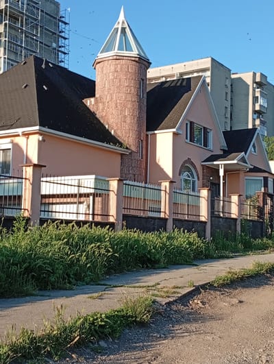 A peach-colored house with a black roof and a cylindrical brick tower with a glass pyramid top is situated in the city of Popovo, Bulgaria. The house is bordered by a black metal fence with stone posts, behind which lush green grass grows. In the background, a tall building under construction with scaffolding and several other residential buildings are visible. The sky is clear and blue, with a few birds in flight. A cracked concrete sidewalk runs in front of the house, leading to a dirt road. The scene is bathed in the warm light of the sun.