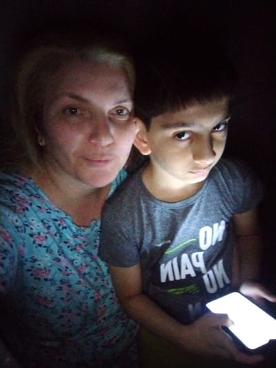 In Al Mas'oudi, United Arab Emirates, a woman and a boy are shown in a dimly lit setting. The woman, positioned on the left, has blonde hair and is wearing a floral patterned shirt. To her right, the boy, with dark hair, is wearing a grey t-shirt with white lettering. He is holding a brightly lit smartphone, which illuminates part of the scene. The background is dark, suggesting an indoor environment at night.