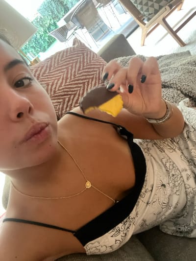 An individual is depicted indoors, reclining on a sofa, in Lordelo do Ouro e Massarelos, Portugal. The subject, a female, is visible from the chest up, positioned on the left side of the frame, gazing towards the camera with slightly pursed lips. She wears a black spaghetti strap top, a light-colored garment with a dark floral pattern, and a delicate gold-colored necklace with small circular pendants. Her right hand, featuring dark nail polish and multiple metallic bracelets on the wrist, holds a partially consumed, yellow-colored confection with a dark chocolate coating. Behind the individual, a reddish-brown and white chevron-patterned cushion and a grey textured throw blanket are visible. The background reveals a brightly lit outdoor area through a large window, featuring lush green foliage, outdoor wicker chairs, and a table, consistent with a patio or garden setting.