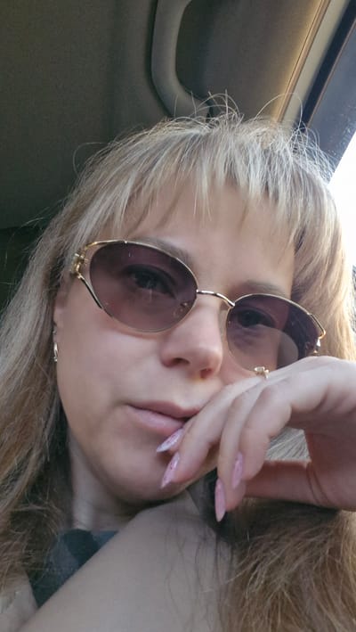 The image shows a blonde woman with long, styled nails, wearing sunglasses with a gold frame, looking directly at the camera. The background suggests she is inside a car, evidenced by the car interior and a handle visible in the upper right corner. The location in the city of Otchor, Russia is not discernible from this image.