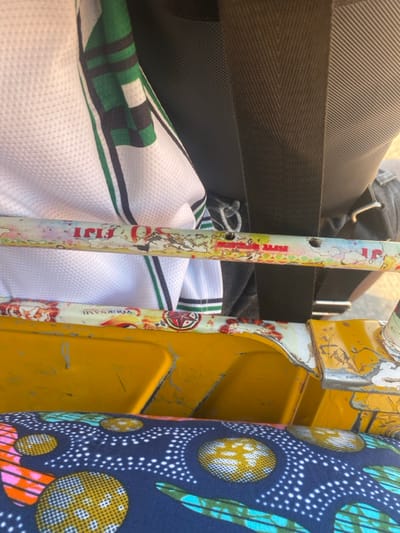 The image shows a cluttered space. In the background, a dark gray backpack with a black strap is visible. In the foreground, a white and green sports jersey is partially visible. A white pole with colorful, peeling graphics and two holes runs vertically. To the right of the pole, a yellow, metal structure with chipping paint stands. A blue fabric with a pattern of circles and white dots is seen on the far right, adding to the overall chaotic impression.