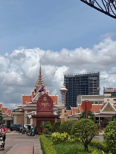 This image depicts the border crossing into Bavet Quarter, Cambodia. In the foreground, a woman pushes a baby in a stroller on a paved walkway. Behind them, a complex of buildings with traditional Cambodian architecture, featuring red tiled roofs and ornate details, is visible. A flag of Cambodia flies from a spire on one of the buildings. To the right, a modern, unfinished high-rise building stands out against the sky, indicating ongoing construction in the area. Several vehicles, including cars and motorcycles, are parked in front of the buildings. Lush green bushes and trees line the foreground and areas around the buildings, suggesting a well-maintained landscape. The sky is partly cloudy, with dramatic white clouds against a light blue backdrop.