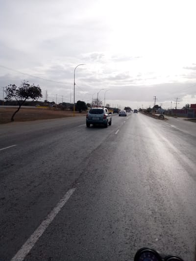The image shows a wide, multi-lane asphalt road in what appears to be La Cruz del Pastel, Venezuela. The road is wet, possibly from recent rain. Several cars are driving in the same direction, including a light blue hatchback directly ahead. A tree is visible on the left side of the road. Utility poles and wires line the roadside. The sky is overcast. A speedometer is visible at the very bottom of the image, suggesting it was taken from a moving vehicle.