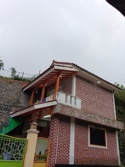 A two-story brick building under a cloudy sky stands on a hillside. The structure has a red tiled roof with white trim and exposed wooden rafters. A small balcony is present on the second story. A green fence and gate are visible in the foreground. The surrounding area features stone retaining walls and greenery. A window is visible on the side of the building, which is painted black.