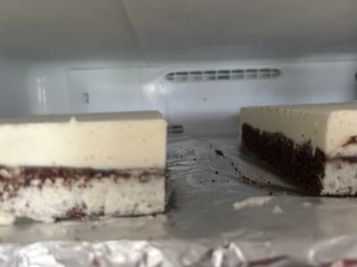 Two rectangular slices of layered dessert, likely cake or cheesecake, are positioned on a sheet of aluminum foil inside a refrigerator. The dessert features a dark brown bottom layer and a thick, creamy white top layer. Crumbs from the dark layer are scattered on the foil and the white topping. The background shows the interior of a refrigerator, including a vent with horizontal slats. The image appears to have been taken in a residential setting in Richmond Hill, Canada.