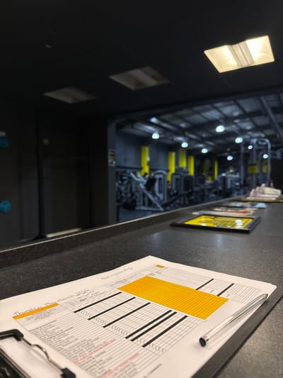 An interior image depicts a gym or fitness facility in Tunbridge Wells, United Kingdom, viewed from behind a dark, textured counter. In the foreground, a white paper schedule, dated "Thursday 19th March 202X," rests on a black clipboard with a silver pen. The schedule displays a grid format with a large yellow block and rows containing black horizontal lines, along with handwritten notes including "Have a great day!" and names like "Liv" and "Kim." Beyond the counter, the gym area is visible, featuring dark walls and ceiling, contrasting with bright yellow vertical support pillars. Numerous black and metallic exercise machines are arranged on the floor. Overhead lighting consists of multiple circular, recessed white lights and square ceiling lights. A dark wall to the left features several blue, circular objects resembling climbing holds. No individuals are clearly depicted.