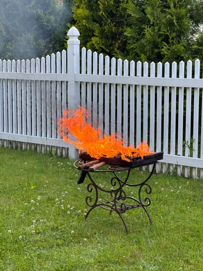 In a grassy yard in Gvardeysk, Russia, a fire burns intensely in a metal barbecue grill. The grill, a dark, rectangular metal box with a decorative metal stand, sits on the green grass dotted with small white flowers. Flames, a vibrant orange and yellow, erupt from the grill, reaching upwards and to the right. A wispy plume of gray smoke rises from the fire and drifts leftward, partially obscuring a white picket fence in the background. Behind the fence, lush green trees form a dense backdrop. The scene is captured outdoors, suggesting a daytime setting.